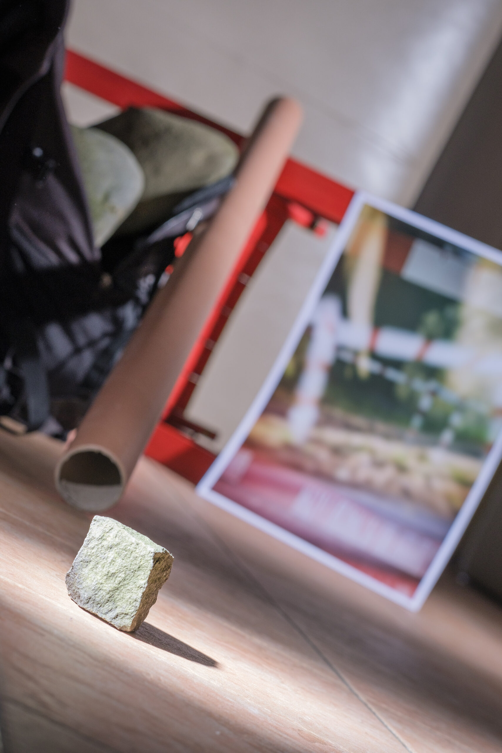 Rucksack mit schweren Steinen im Hintergrund, im Vordergrund angeblitzter Pflasterstein, ein Bild mit rot-weißer Absperrung, ein umgefallener Stuhl und ein Rohr aus Pappe ragt von hinten nach vorne