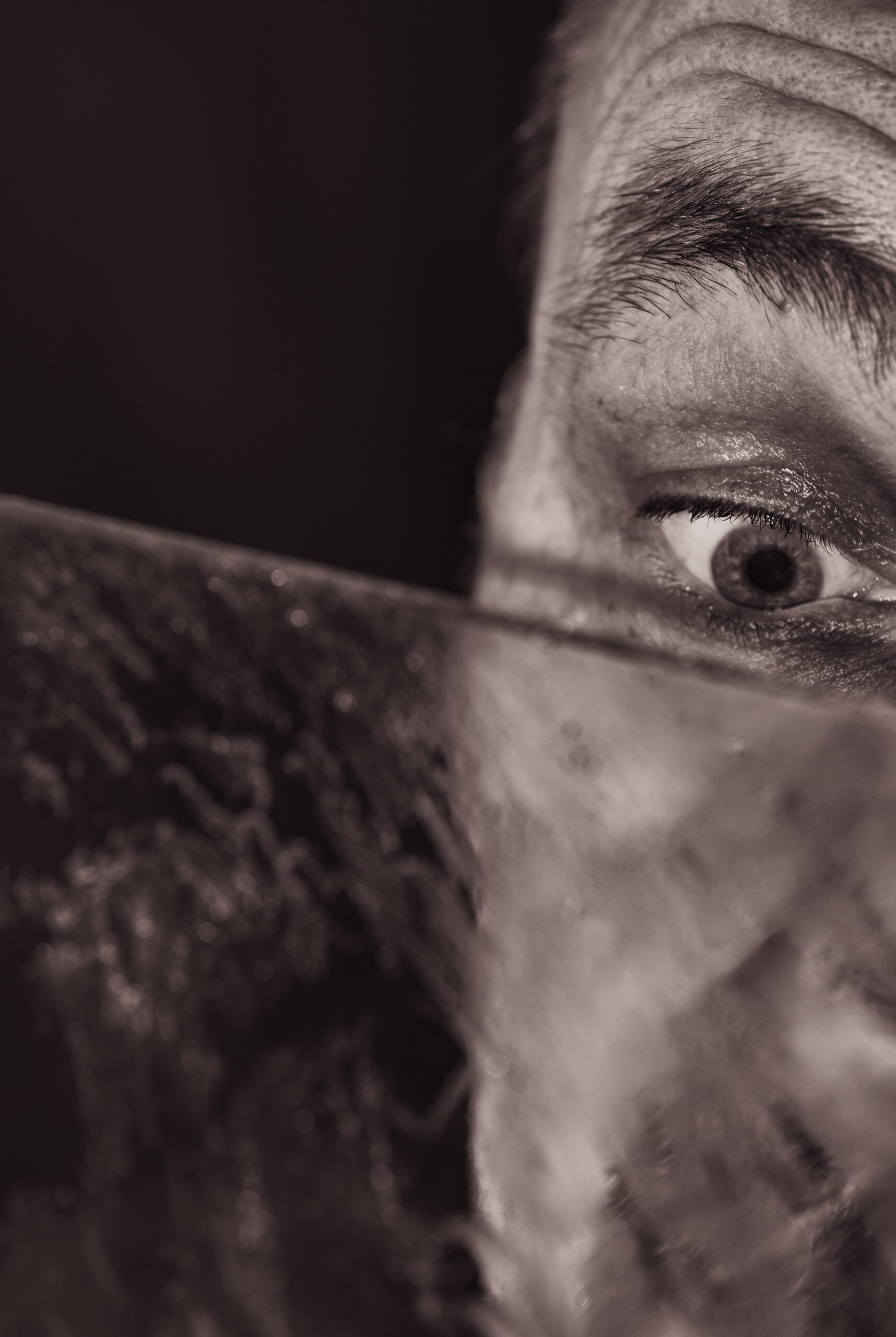 Nahaufnahme einer rechten Gesichtshälfte, der Mann schaut auf den Rand einer schmierigen Scheibe, der Schatten des Randes läuft unter. seinem Auge, entsättigt, sepia
