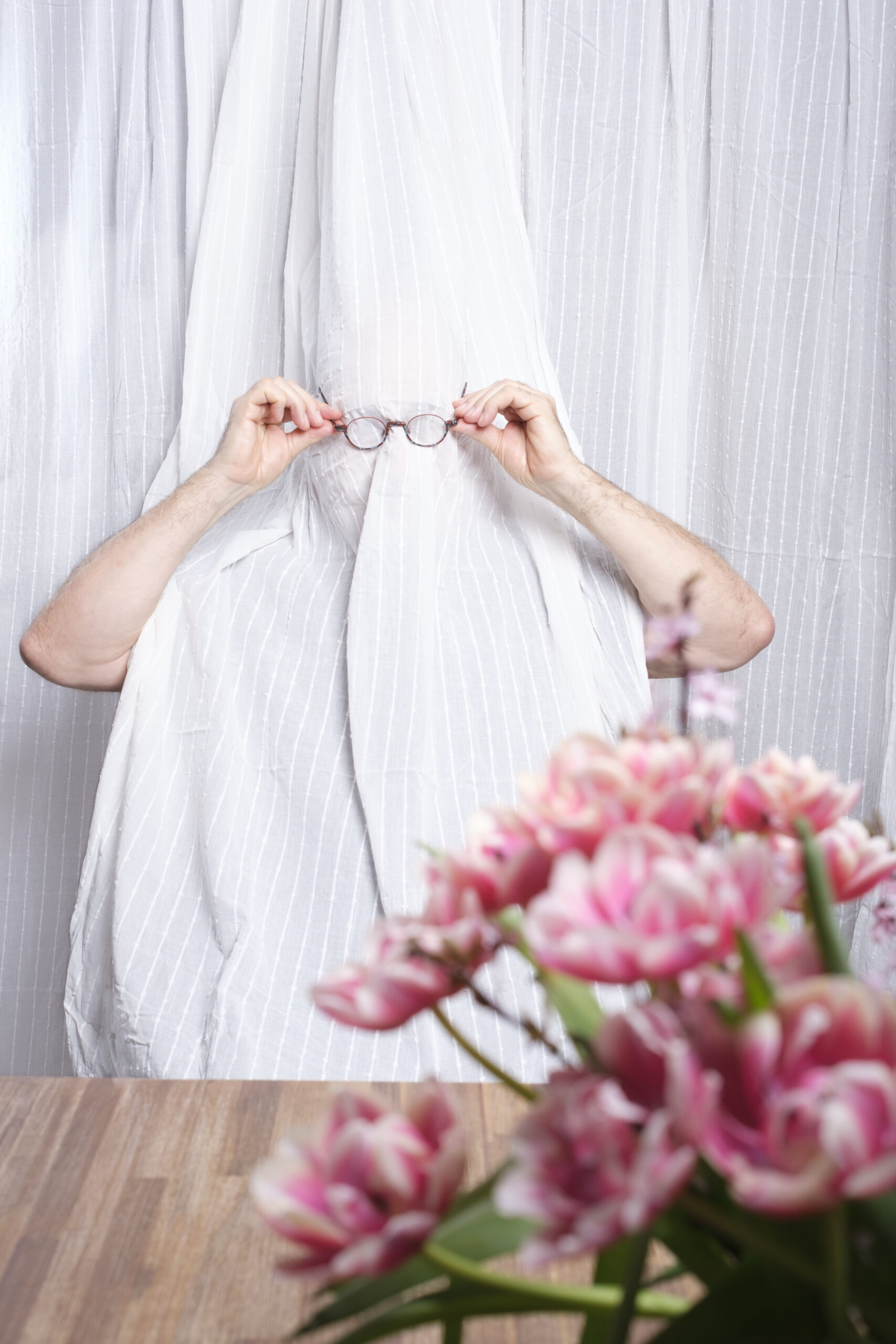 Portrait eines Mannes, sitzend hinter einem Vorhang, nur die Arme sind zu sehen, mit beiden Händen setzt der Mann sich eine Brille auf, im Vordergrund rechts eine Vase mit gefüllten Tulpen in pink