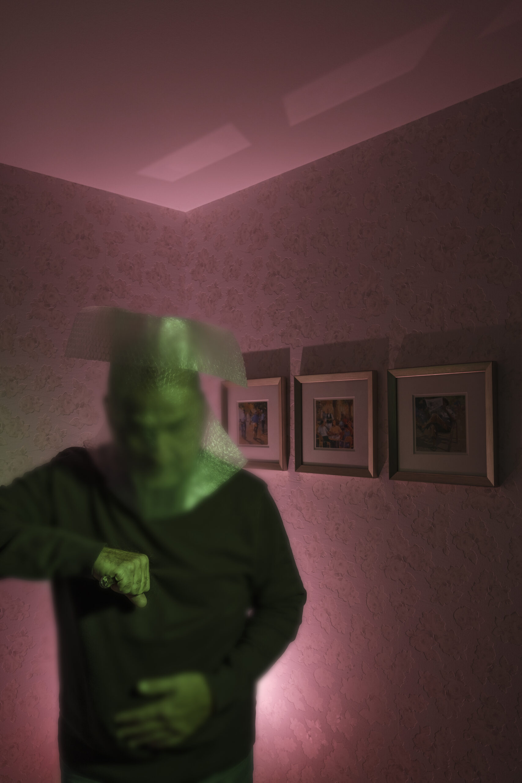 Mann im Vordergrund, teils scharf, teils unscharf abgebildet mit Hand in Richtung Brustkorb, Kopf verfremdet, im Hintergrund eine Wand mit drei Bildern, Licht hinter dem Mann, Schatten an der Raumdecke