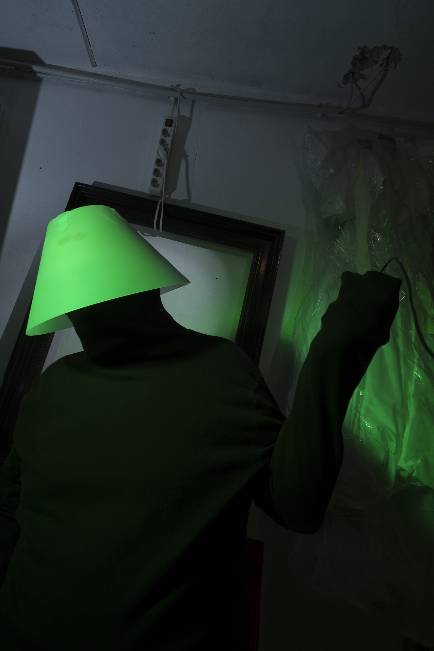 Mann in schwarz mit grünem Lampenschirm, der den Kopf verdeckt, hält eine Fernbedienung in der Hand, im Hintergrund ein schief hängender leerer Bilderrahmen und ein Brautkleid an der Wand hängend