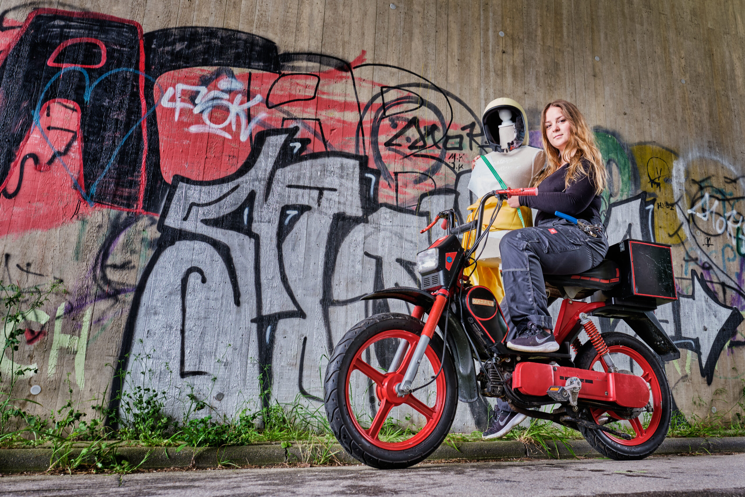 Work-Life-Balance Part One Frau mit Schneiderpuppe und Lochzange im Arbeitsoverall auf Moped vor Graffitti